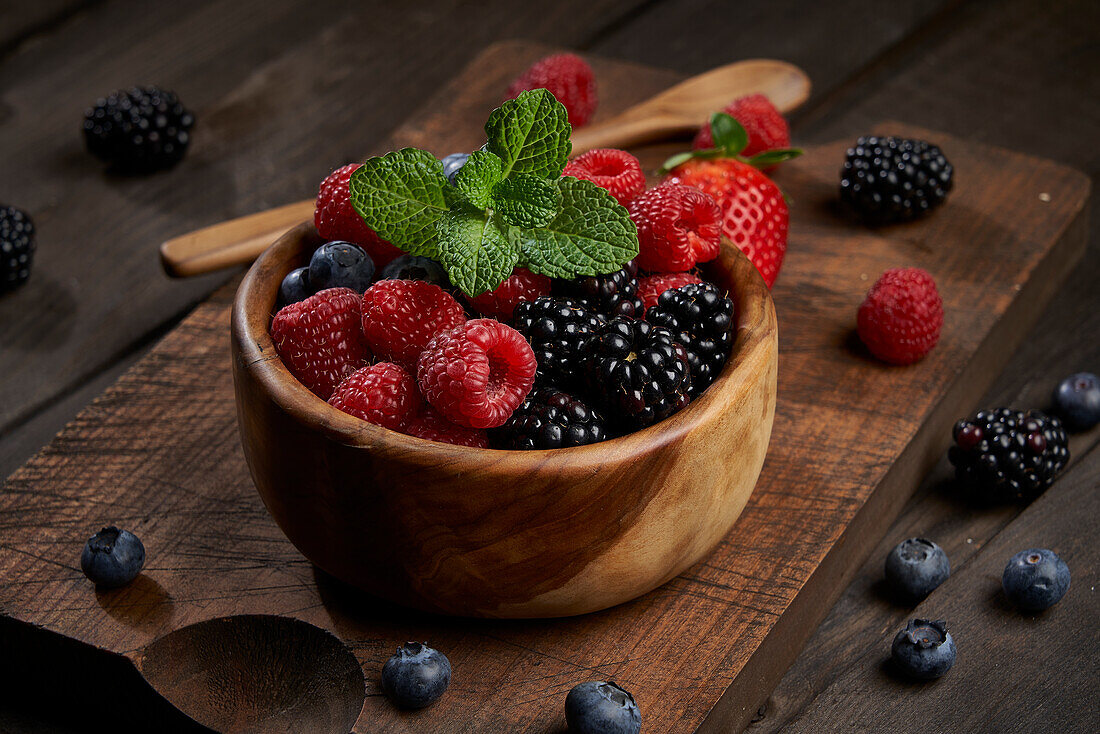 Von oben Schale mit verschiedenen reifen Beeren und Minzblättern auf hölzernem Schneidebrett in heller Küche