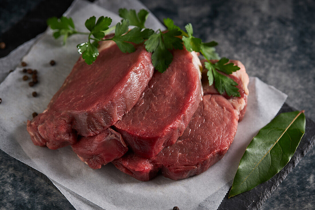 From above of fresh uncooked beef fillet with green parsley placed on slate board with parchment paper in kitchen