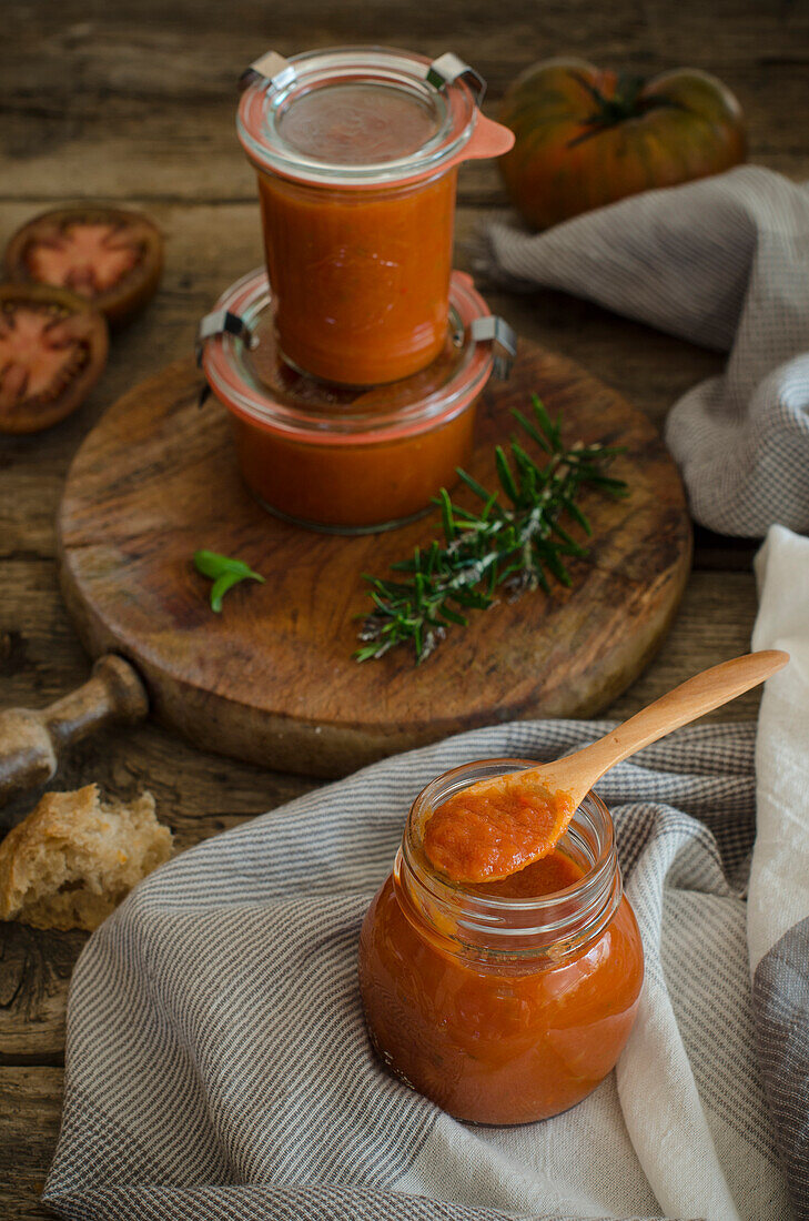 Full glass jars with homemade natural fried tomato sauce placed on cutting board board near with fresh green rosemary and basil leaves placed on rustic wooden table