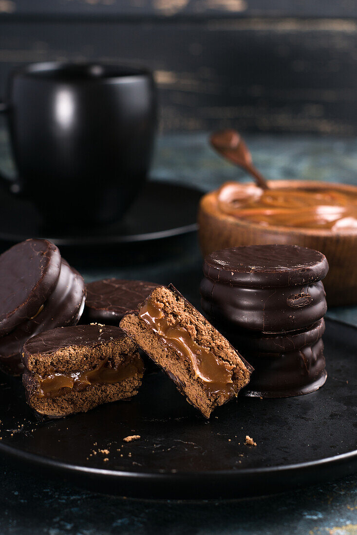 Schokoladen-Alfajores auf einem Teller und Dulce de Leche in einer Schüssel neben einer Tasse mit heißer Schokolade