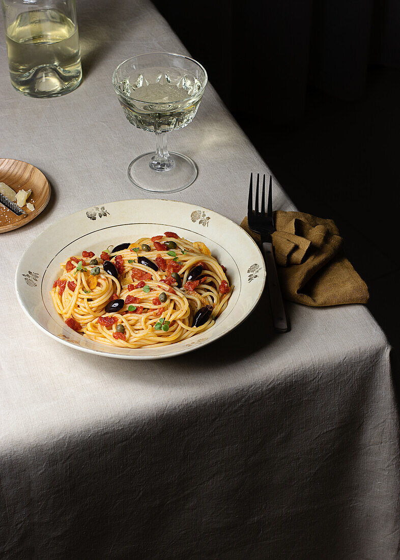 Spaghetti alla Puttanesca, serviert mit einem Glas Weißwein auf einem Tisch mit Serviette