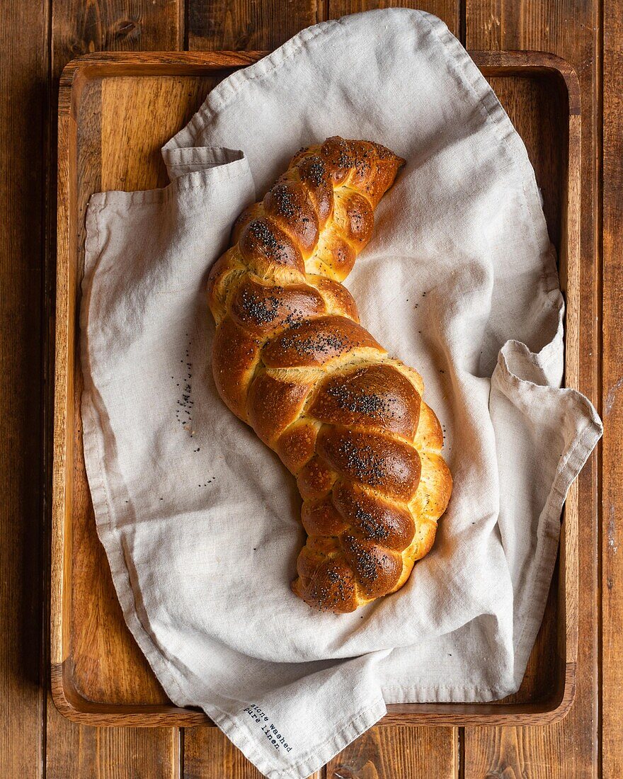 From above of served golden brown bread braid challah with poppy seeds on white napkin in tray