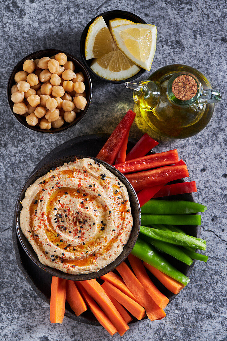 Top view of delicious hummus and slices of assorted vegetables served near bottle of oil and bowls of chickpea and lemon pieces on gray marble table