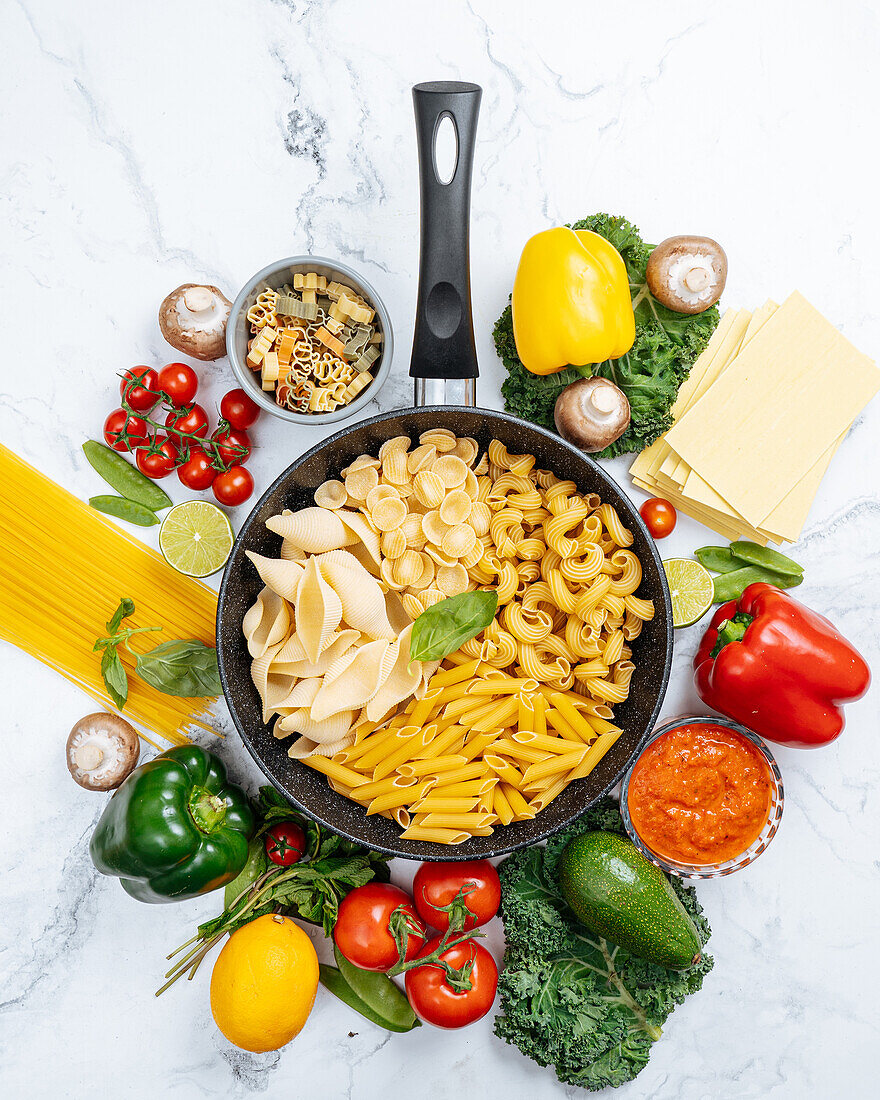 From above of frying pan with various uncooked pasta surrounded with assorted raw vegetables on marble table in light kitchen