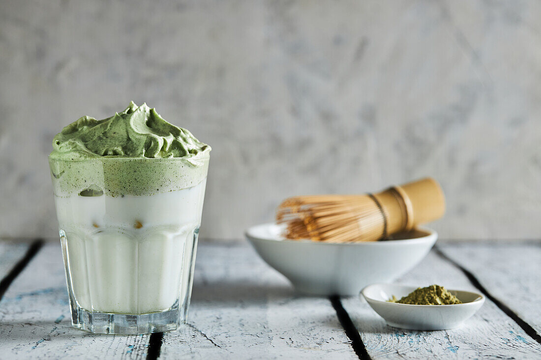 Glas frischer Dalgona-Kaffee mit Matcha und japanisches Teezeremonie-Zubehör, bestehend aus einem Bambusbesen namens Chasen, auf einem Holztisch