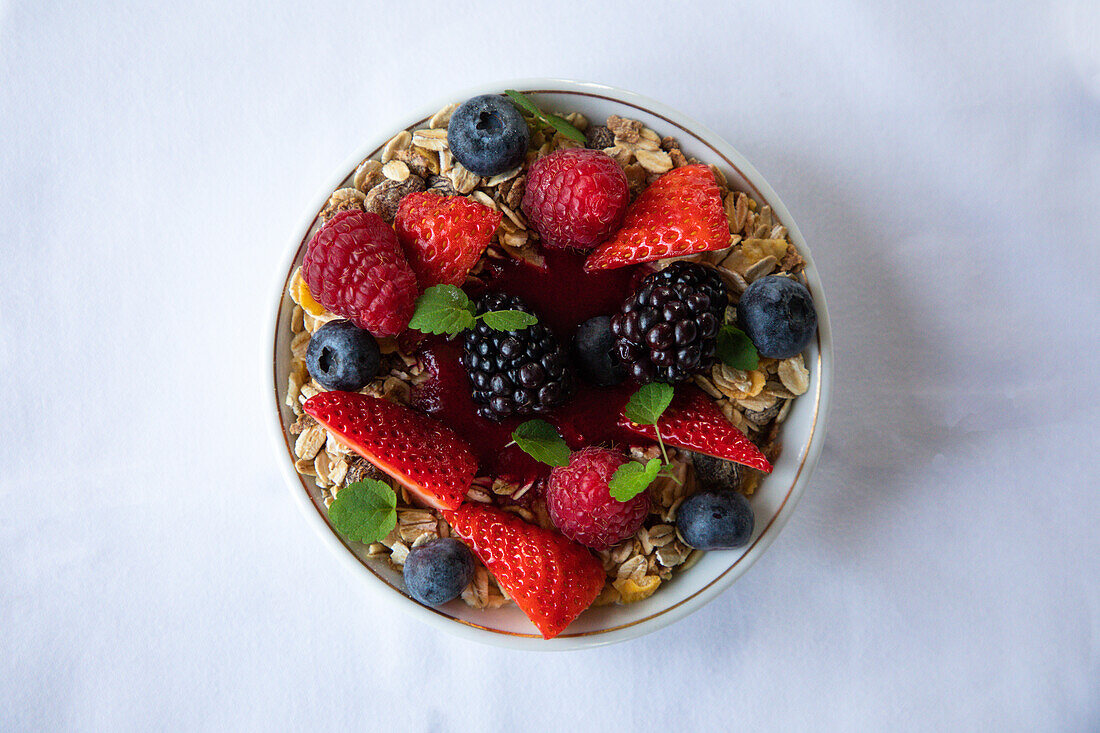 Top view of appetizing muesli bowl decorated with various fresh berries and mint leaves served on white table in morning