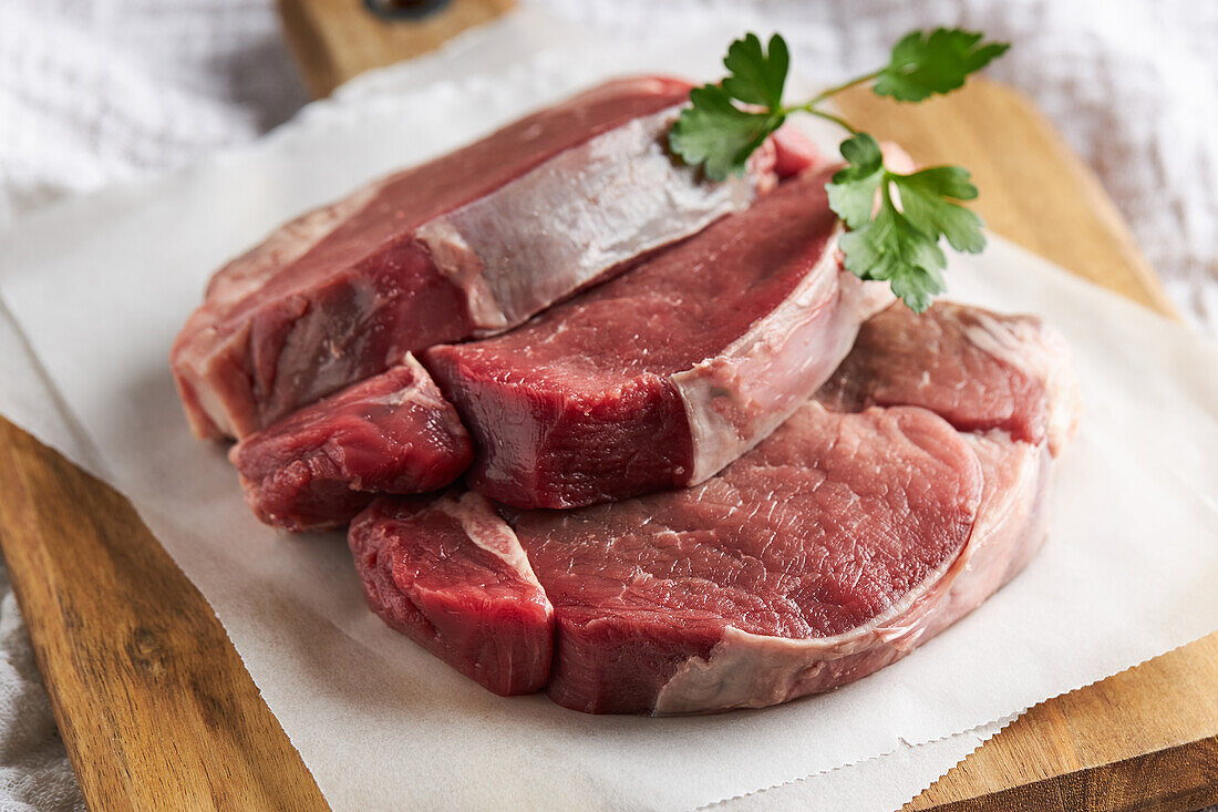 Von oben Scheiben von frischem, ungekochtem Rinderfilet mit grüner Petersilie auf einem hölzernen Schneidebrett mit Pergamentpapier in der Küche