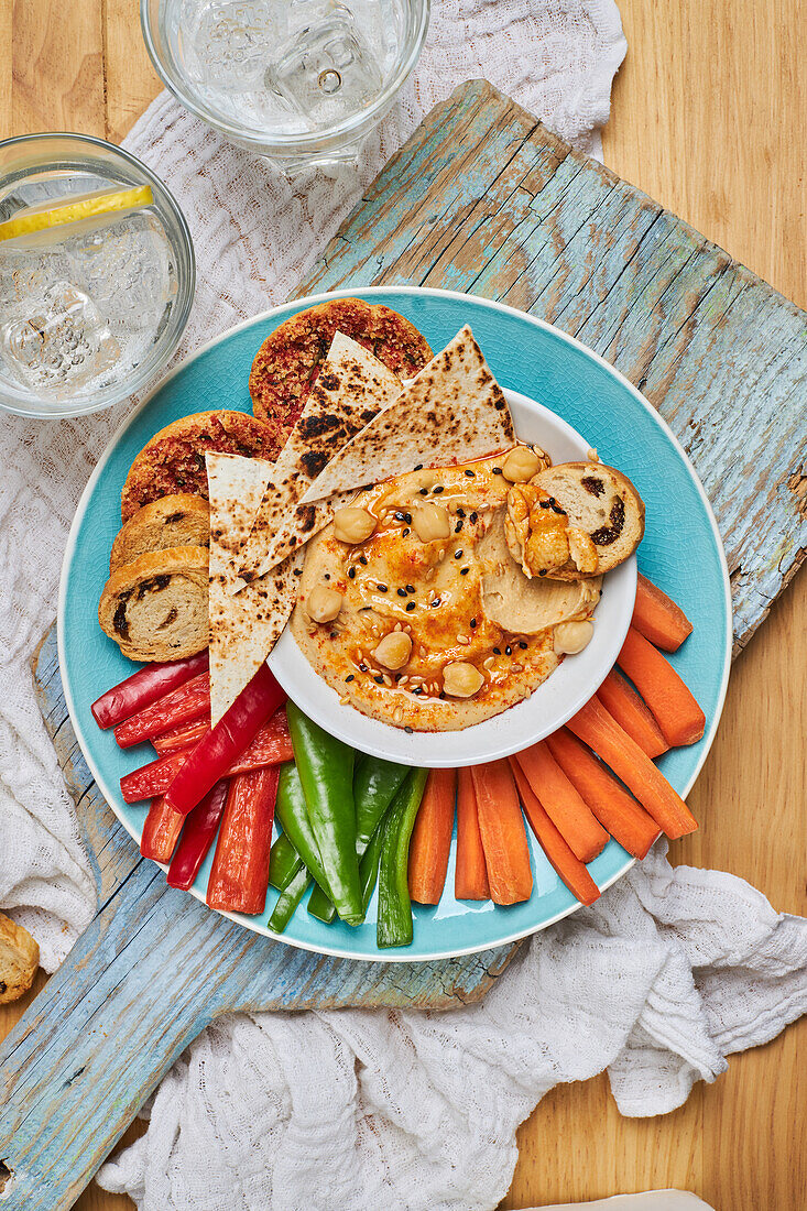 Draufsicht auf leckeren Hummus mit Tortilla-Chips, serviert auf einem Teller mit Croutons und verschiedenen Gemüsesorten auf einem rustikalen Schneidebrett neben Gläsern mit kalter Limonade auf einem Holztisch mit Serviette