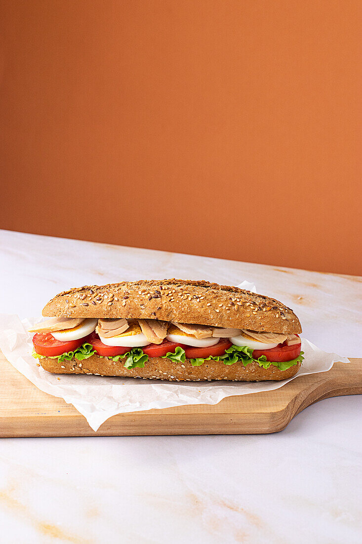 Appetizing sandwich with hard boiled egg, fresh lettuce, tuna and tomatoes served on baking paper on chopping wooden board on colorful background