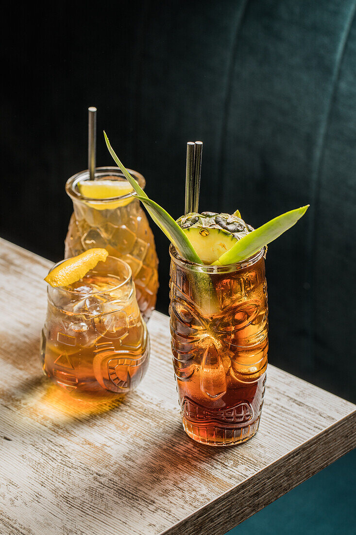 From above of glasses tiki mug filled with alcohol beverage with straw decorated with fruit placed on edge of wooden table couch