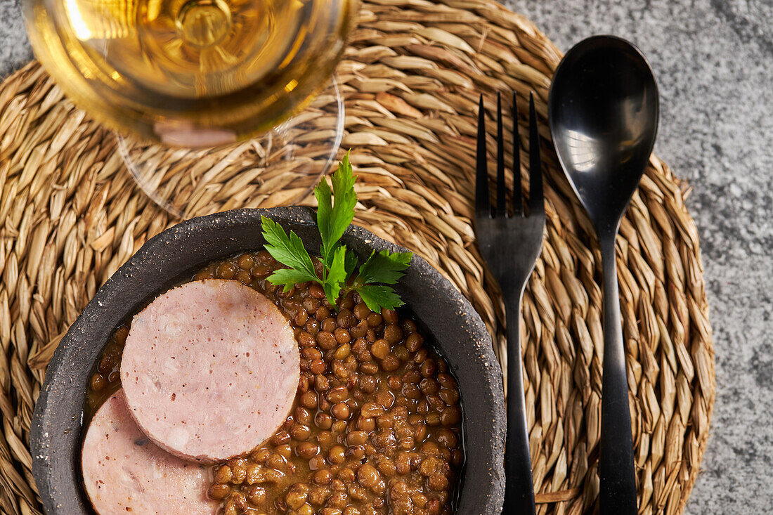 Rustikale Schale mit leckerer Linsensuppe mit Petersilie und Wurstscheiben auf Marmortisch neben Wein und Besteck beim Mittagessen