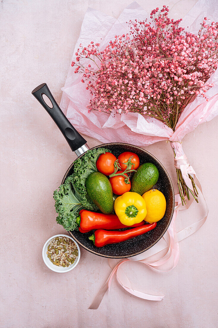 Top view of frying pan with abundance of uncooked vegetables on pink background with bouquet of Gypsophila flowers in paper wrapping