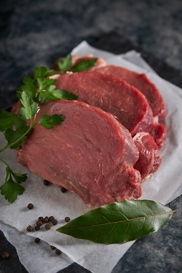 Draufsicht auf Scheiben von frischem, ungekochtem Rinderfilet mit grüner Petersilie auf einer Schiefertafel mit Pergamentpapier in der Küche