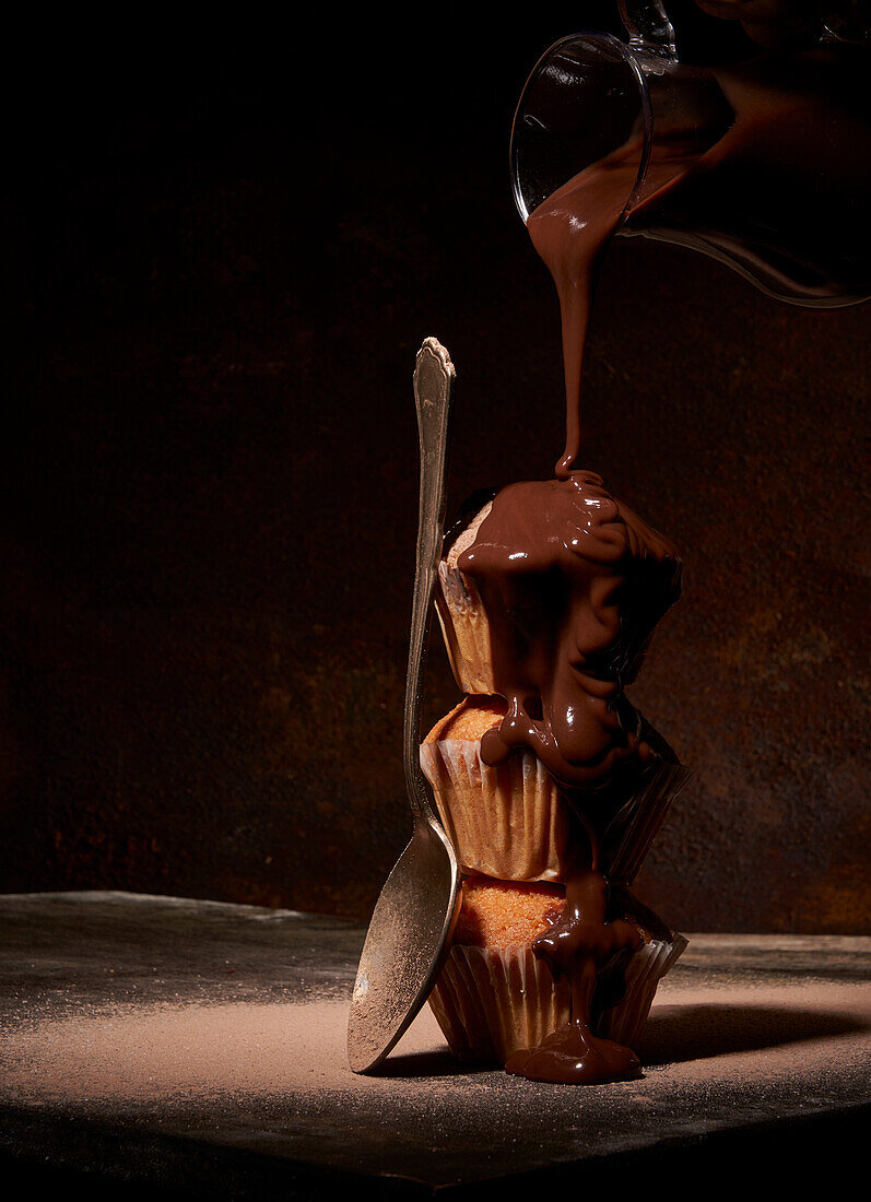 Sweet chocolate topping pouring on tasty naked muffins served on top of each other near spoon on black background in room