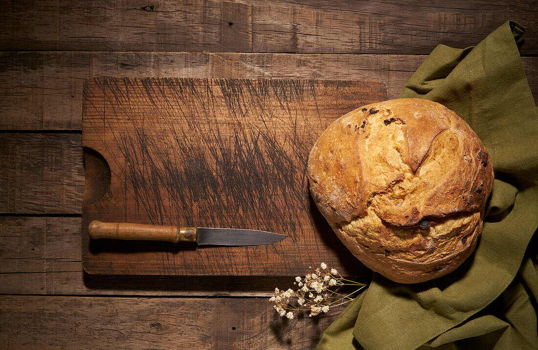 From above of whole round freshly baked artisan bread loaf with crispy crust placed on wooden cutting board with knife on rustic table