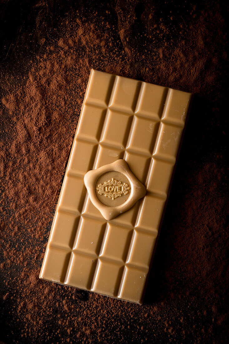 Von oben auf eine ganze Tafel Schokolade mit der Aufschrift "Love", serviert auf schwarzem Hintergrund mit verstreutem Kakaopulver
