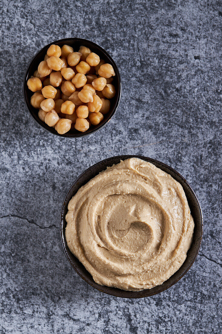 Draufsicht auf Schüsseln mit frischem Hummus und Kichererbsen auf einem grauen Marmortisch in der Küche