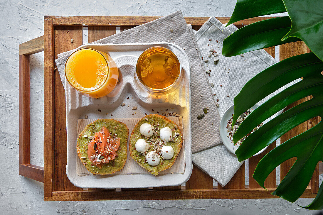From above of delicious avocado toasts with salmon and burrata cheese served on table with glasses of fresh juice and herbal tea placed near exotic Monstera deliciosa plant