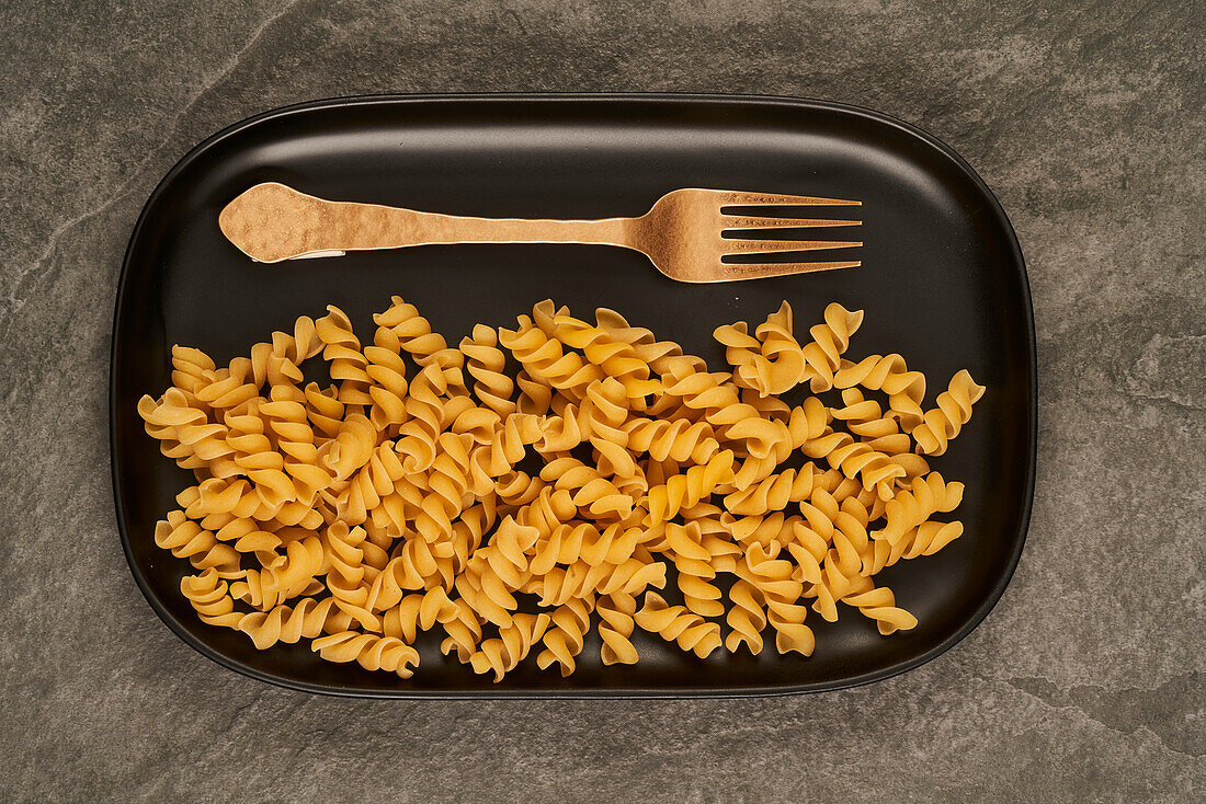 Top view of gold fork placed near uncooked fusilli pasta on tray on table