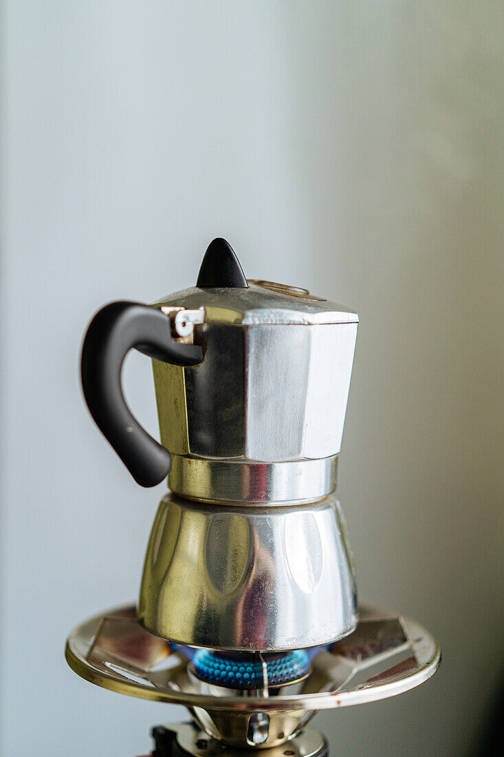 Moderne Metall-Moka-Kanne zum Aufbrühen von Kaffee auf einem rostfreien Gasherd in einer hellen Küche mit Tassen im unscharfen Hintergrund