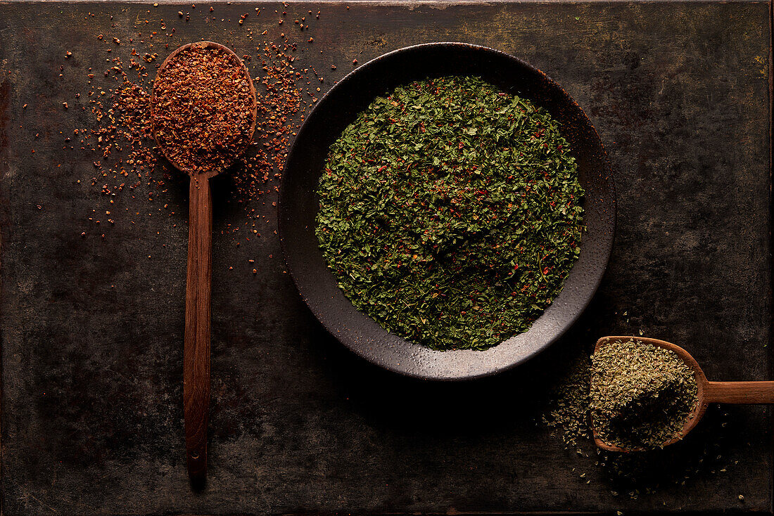 Top view of bowl with green dried herbs and spoon with ground sun dried tomato powder with artificial wooden hand on black background