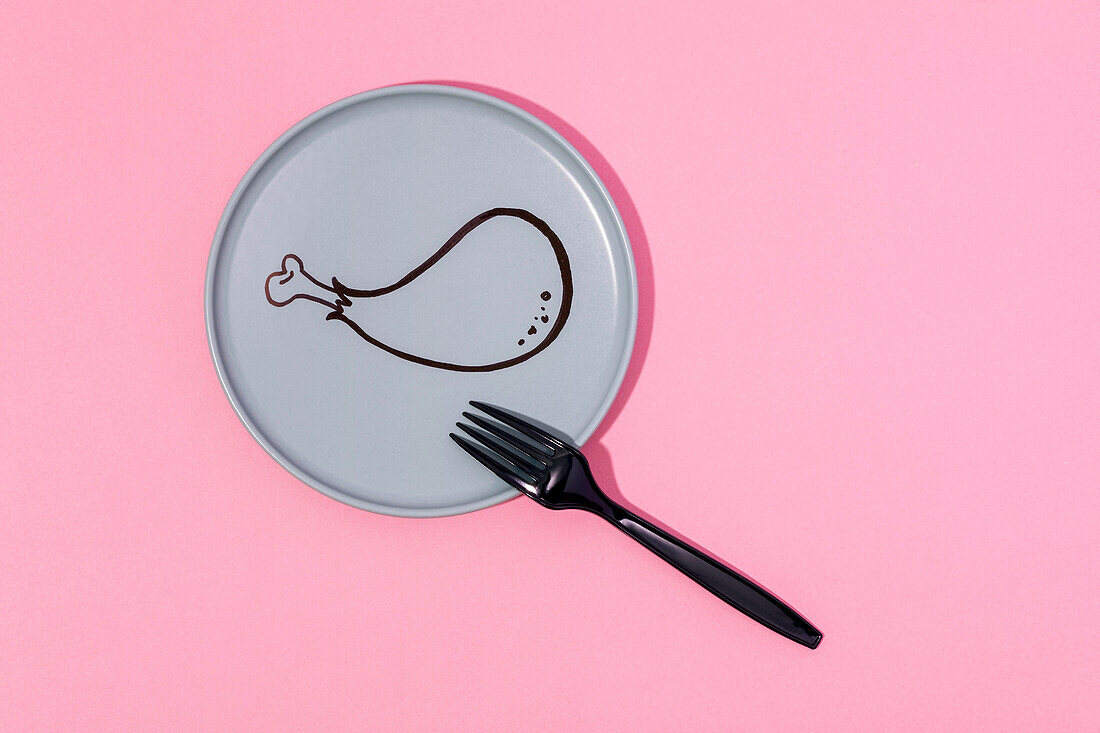 Schwarze Plastikgabel von oben auf rosa Tisch auf grauem Teller mit gemalter Cartoon-Hühnerkeule