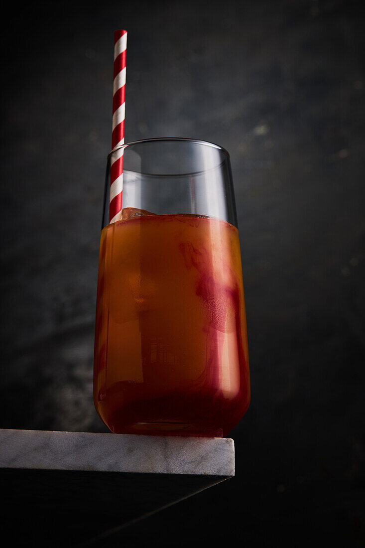 Transparent glass of red alcohol cocktail served with striped straw served on corner of marble table against gray background in studio