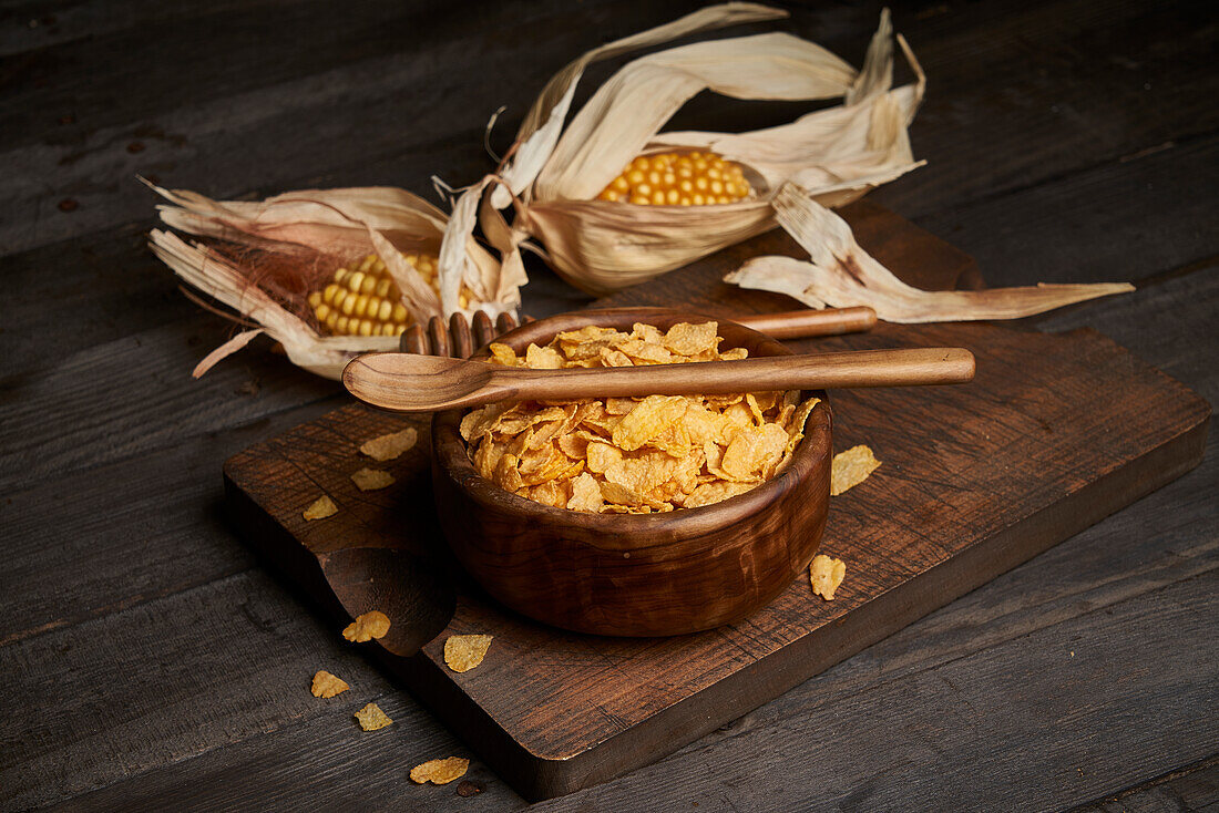 Schüssel mit leckeren süßen Cornflakes mit Löffel auf hölzernen Schneidebrett auf dem Tisch mit Maiskolben in hellen Küche platziert