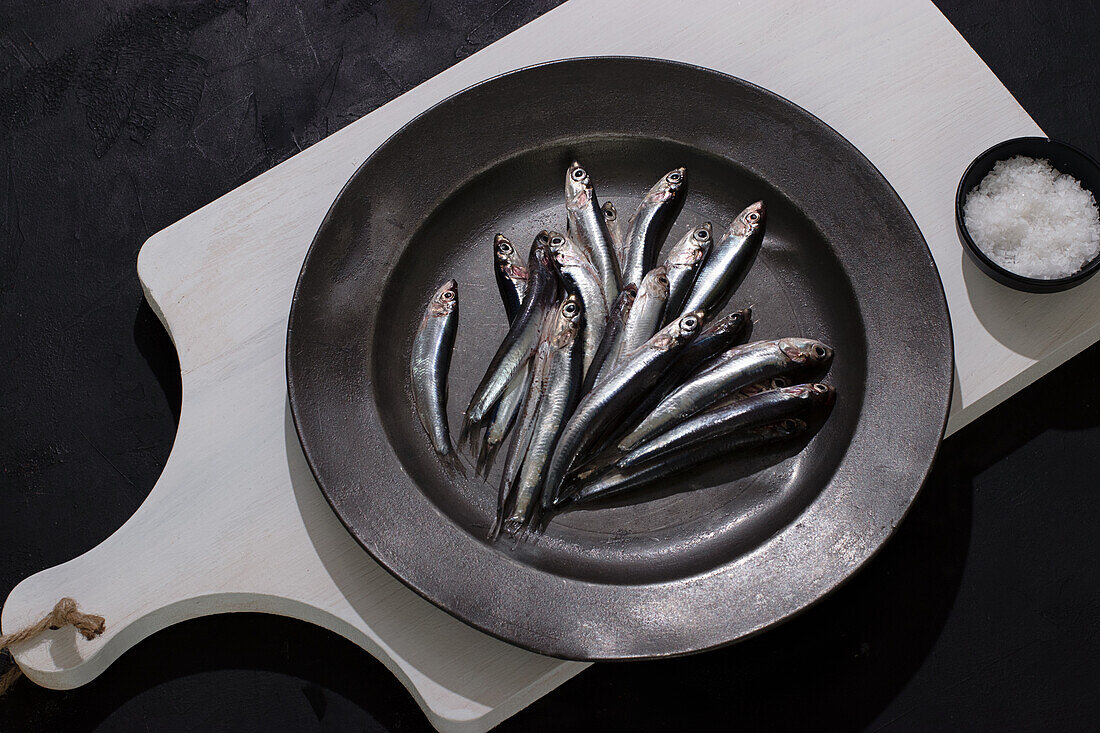 Draufsicht auf rohe kleine Fische in einem Teller auf einer weißen Tafel auf schwarzem Hintergrund