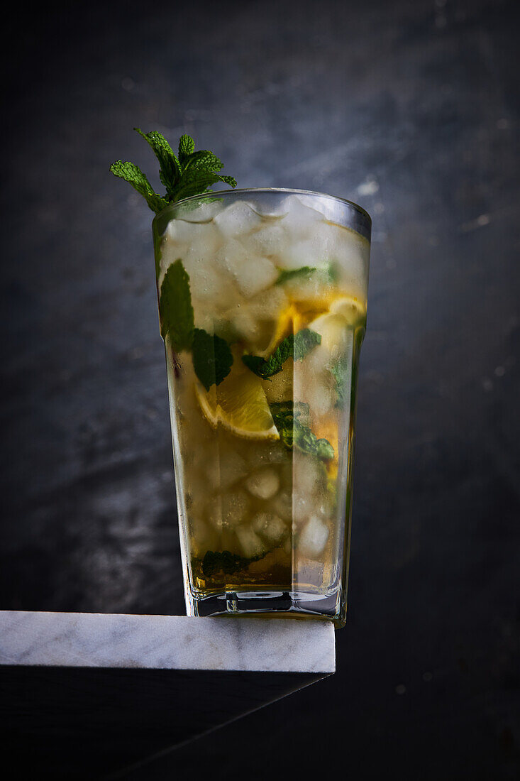Transparentes Glas mit Alkoholcocktail mit Zitronenscheiben und grünen Minzblättern mit Eis, serviert auf einer Tischecke auf grauem Hintergrund