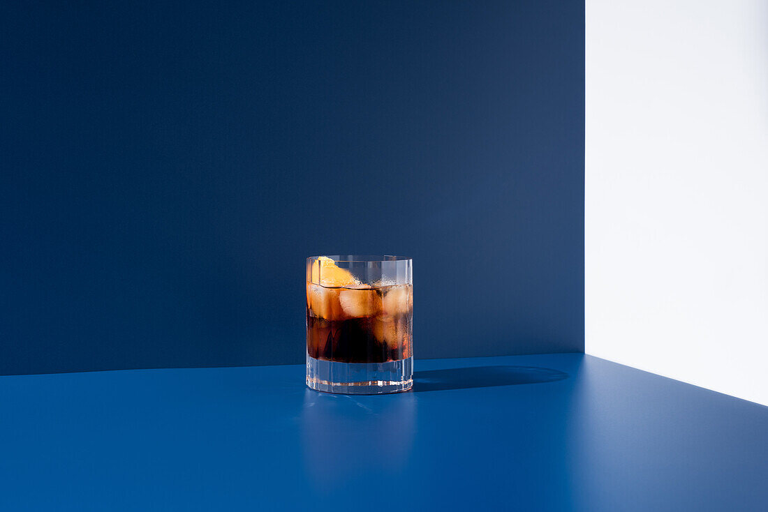 Glass of alcoholic cocktail made with red vermouth served with orange slices in light studio with blue and white wall