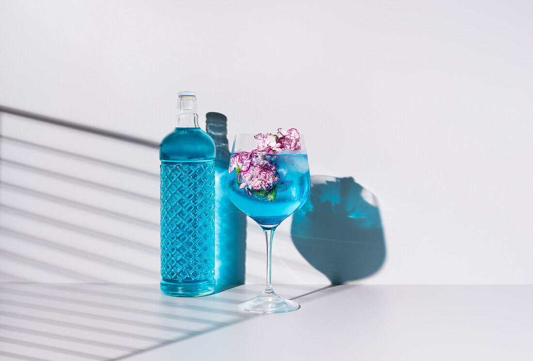 Glass bottle of blue colored liquid placed near glass with refreshing cocktail with ice and flowers on table against white background
