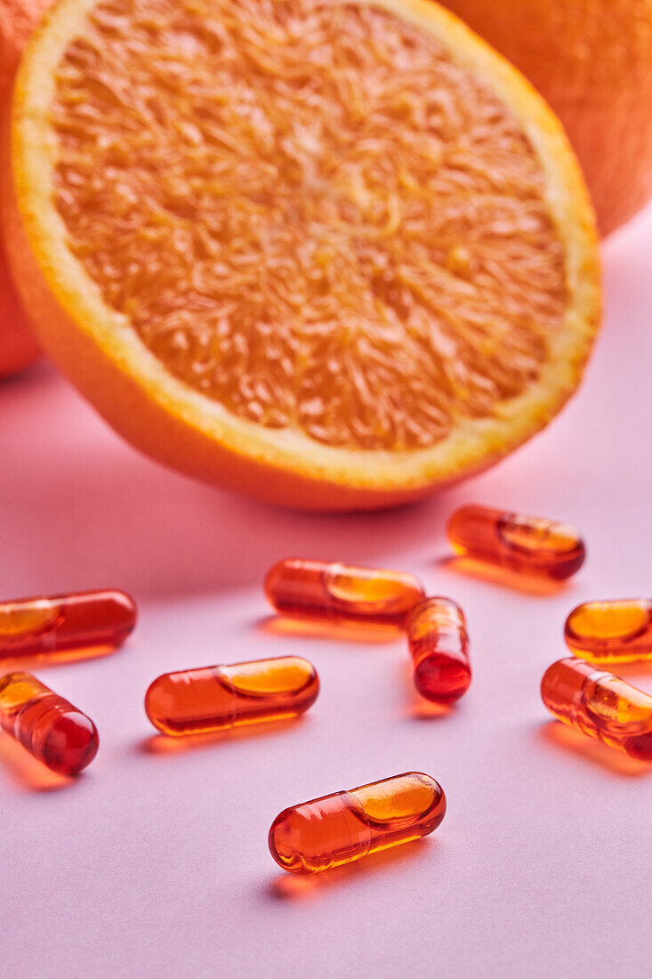 Composition of ripe cut oranges arranged on pink surface near scattered pills in light studio