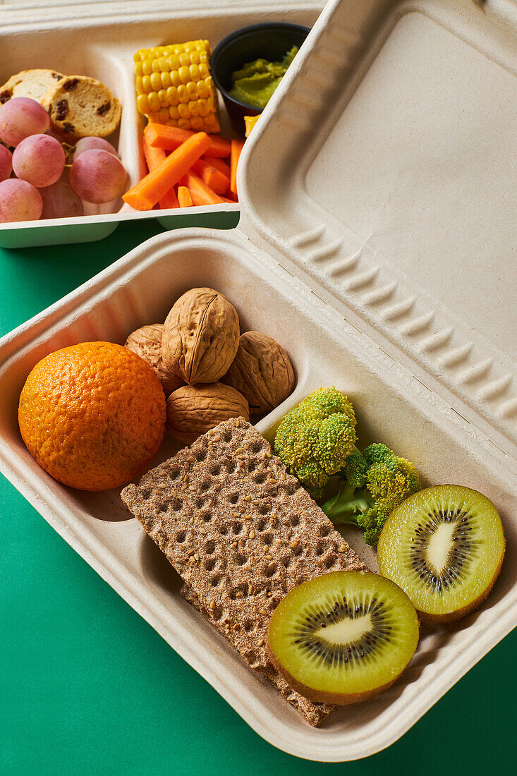 From above of lunch boxes with healthy food including crackers carrot sticks grapes cherry tomatoes with kiwi broccoli walnut and tangerine on green background