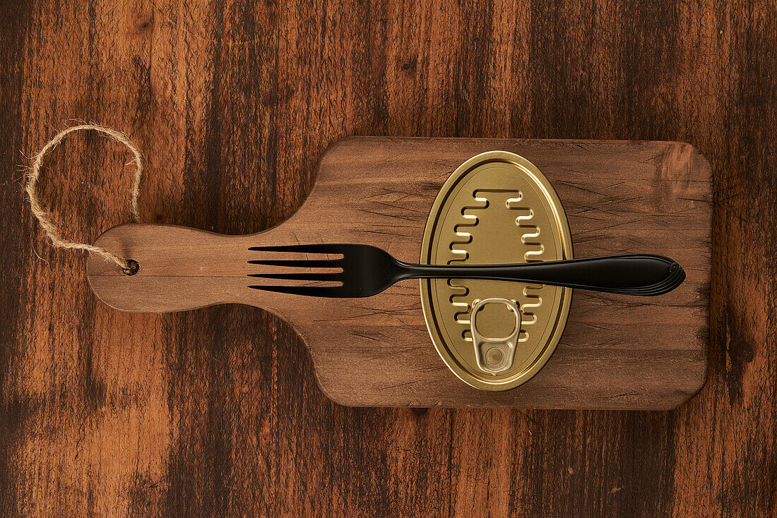 From above scratched chopping board with fork and sealed can with preserved food on rustic lumber table