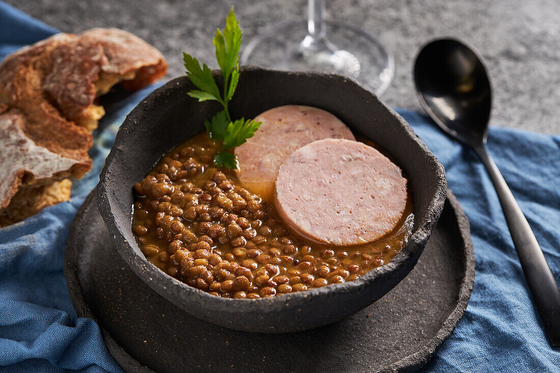 Von oben rustikale Schale mit leckerer Linsensuppe mit Petersilie und Wurstscheiben auf Marmortisch und blauer Serviette neben Brot und Wein beim Mittagessen