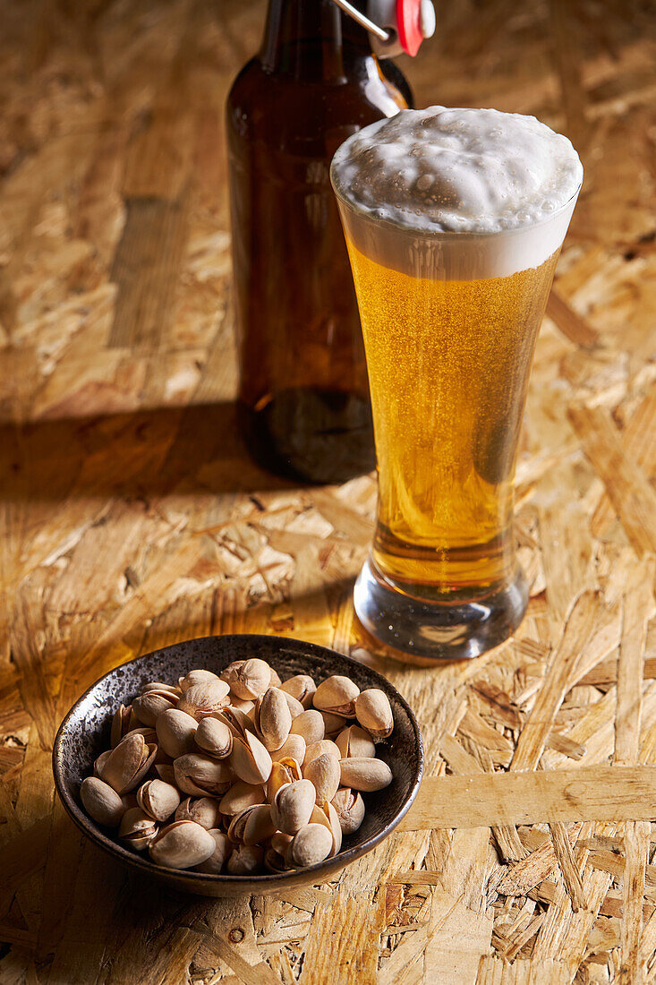 From below glass of fresh cold beer placed on wooden table near bottle and plate with pistachios