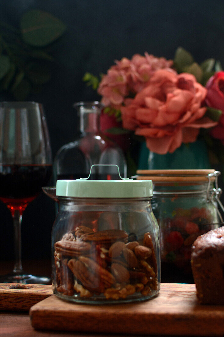 Glass transparent jar filled with nuts placed on wooden cutting board on table with alcohol and blooming flowers in kitchen
