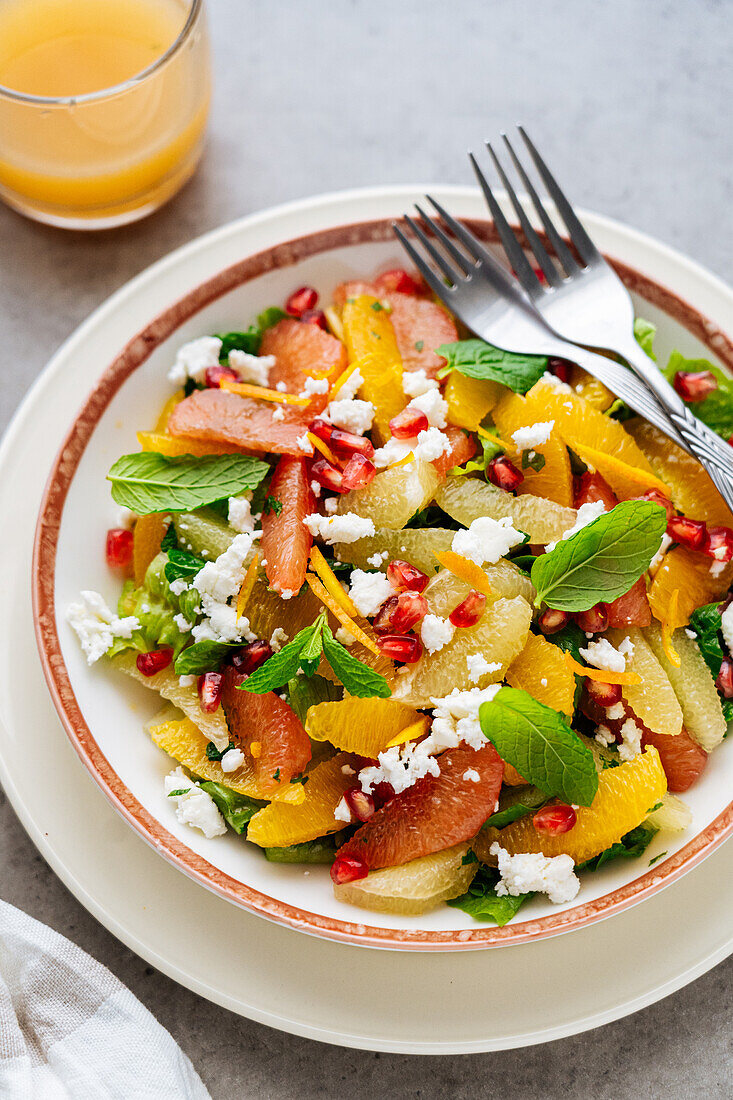 Draufsicht auf traditionellen griechischen Salat mit reifen Tomaten und Feta-Käse, gekrönt von Granatapfelkernen und Minzblättern auf grauem Hintergrund mit Orangensaft