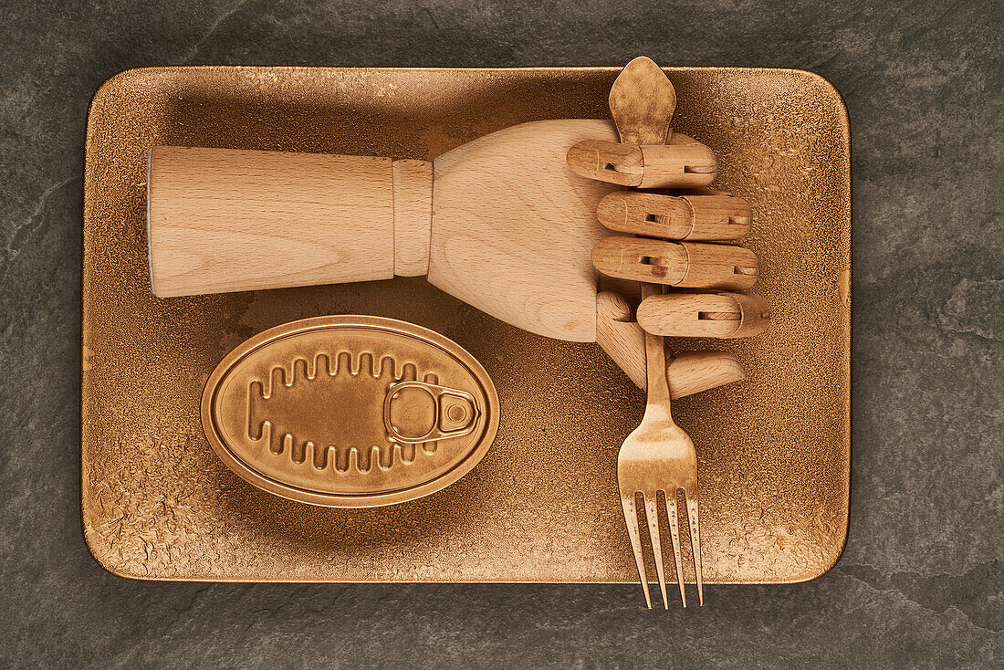 Künstliche Holzhand mit Gabel auf goldenem Tablett neben versiegelter Dose mit Konserven auf dem Tisch