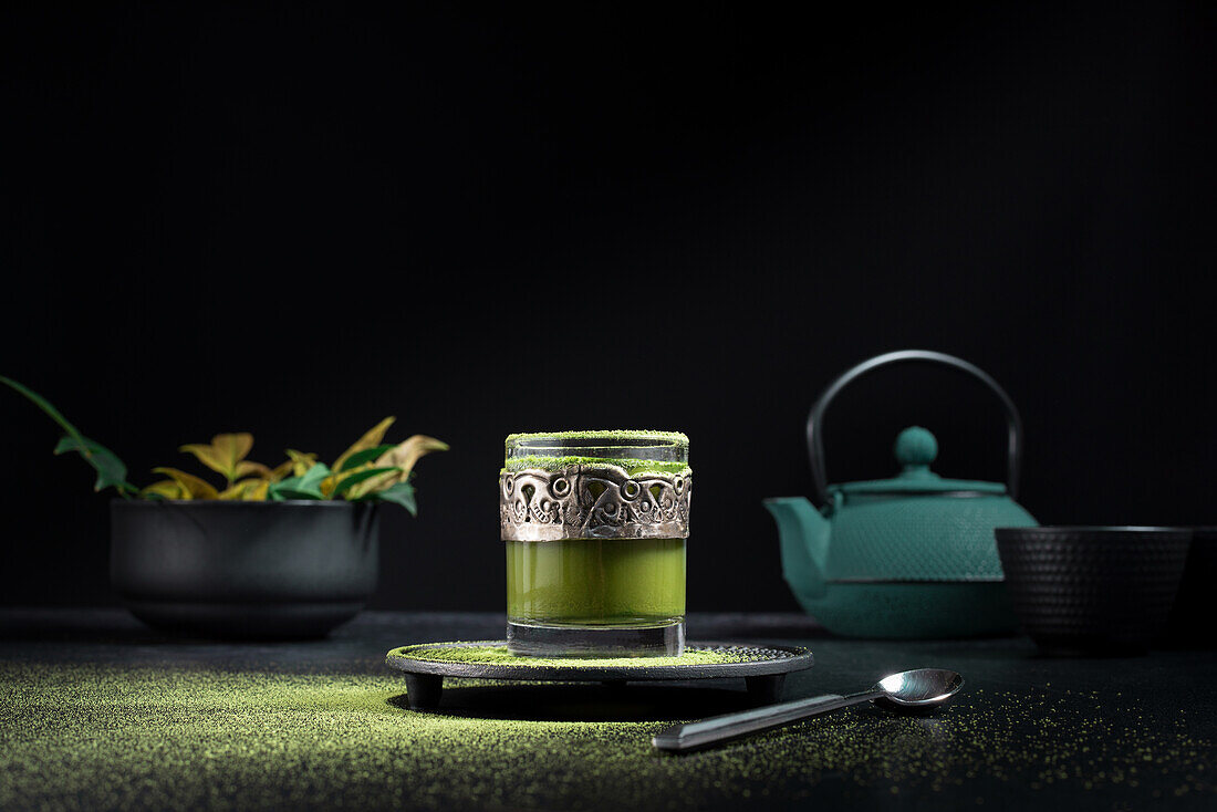 Stillleben Komposition mit traditionellem orientalischen Matcha-Tee in Glastasse mit Metallverzierung auf Tisch mit Keramikschalen und frischen grünen Blättern vor schwarzem Hintergrund