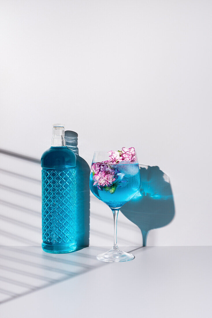 Glasflasche mit blauer Flüssigkeit in der Nähe eines Glases mit einem erfrischenden Cocktail mit Eis und Blumen auf einem Tisch vor weißem Hintergrund