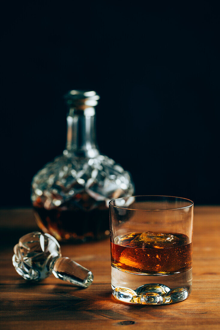 Glass of cold whiskey with ice placed on wooden table near decanter on black background