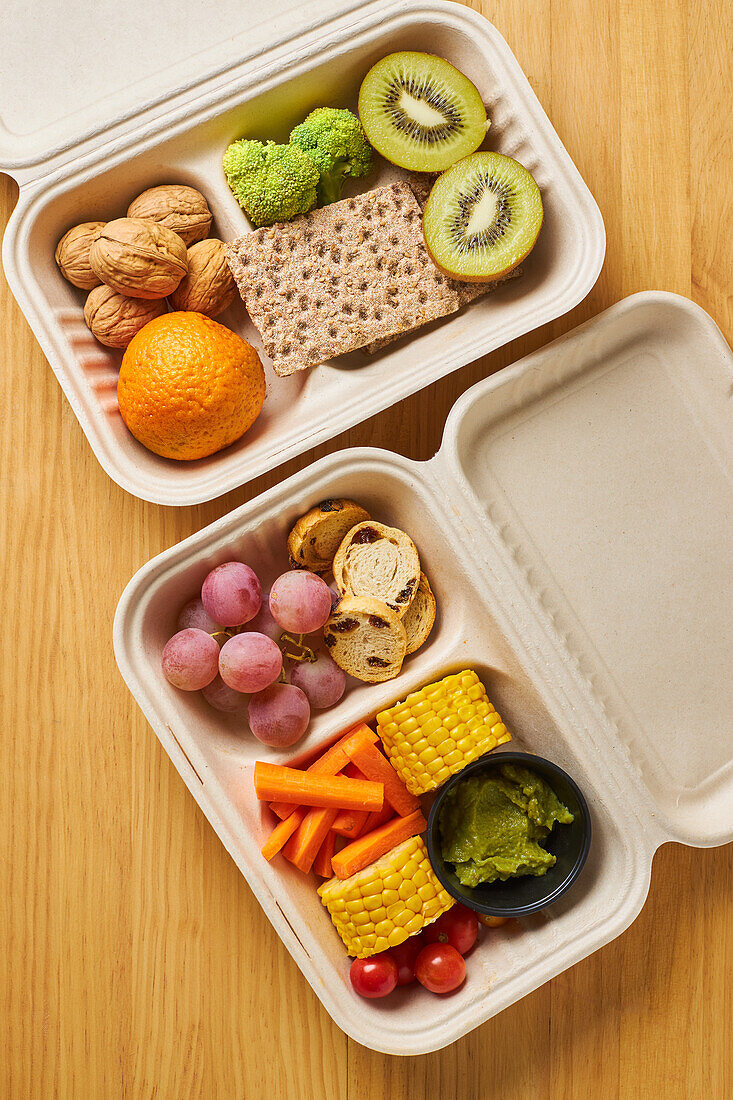 From above of lunch boxes with healthy food including crackers carrot sticks grapes cherry tomatoes with kiwi broccoli walnut and tangerine on yellow background