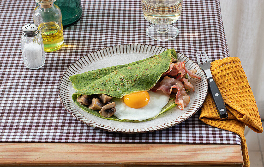 Homemade breakfast of spinach pancakes with bacon, egg and mushrooms served on a white plate with a salt and oil shaker on a checkered tablecloth.