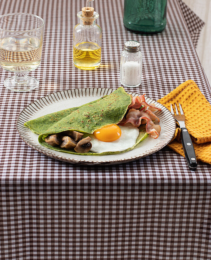 Homemade breakfast of spinach pancakes with bacon, egg and mushrooms served on a white plate with a salt and oil shaker on a checkered tablecloth.