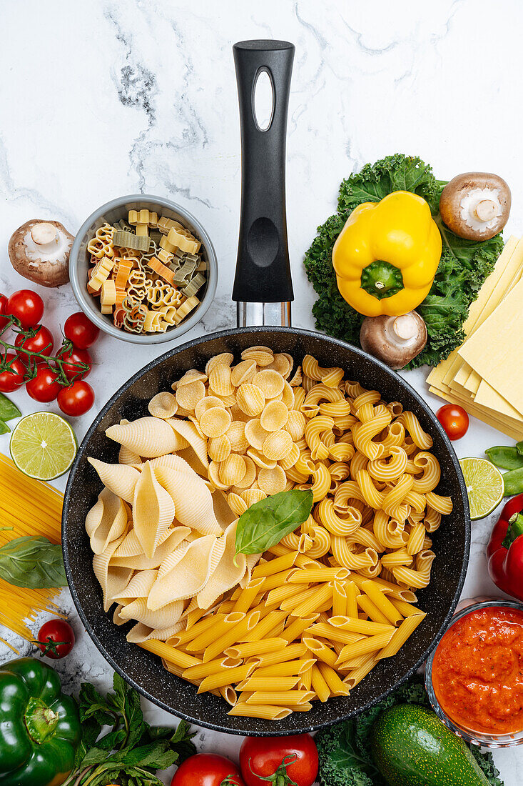 From above of frying pan with various uncooked pasta surrounded with assorted raw vegetables on marble table in light kitchen