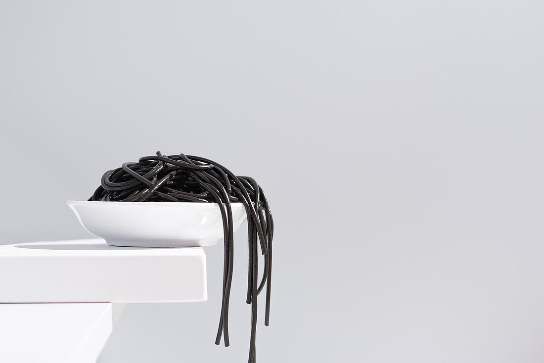 Minimalist studio with black squid ink spaghetti falling out from full ceramic bowl on white table