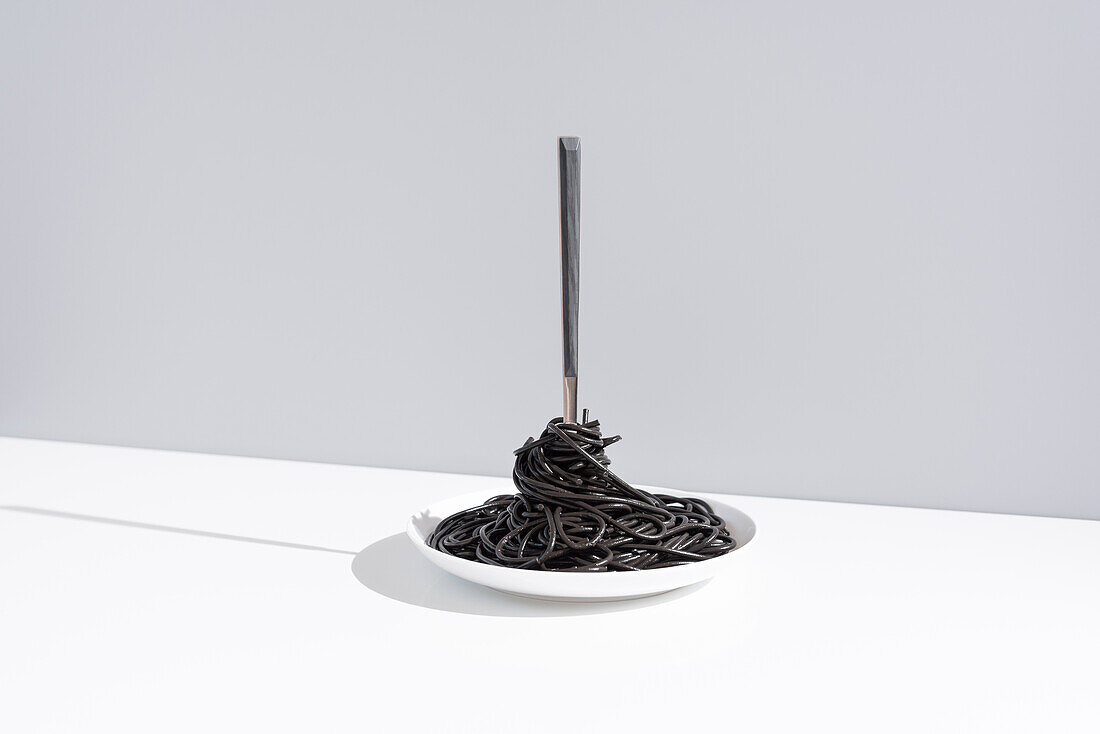 Stainless fork in full bowl of black spaghetti with cuttlefish ink on white table in studio on gray background