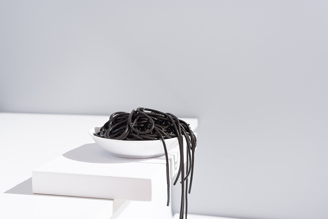 Minimalist studio with black squid ink spaghetti falling out from full ceramic bowl on white table